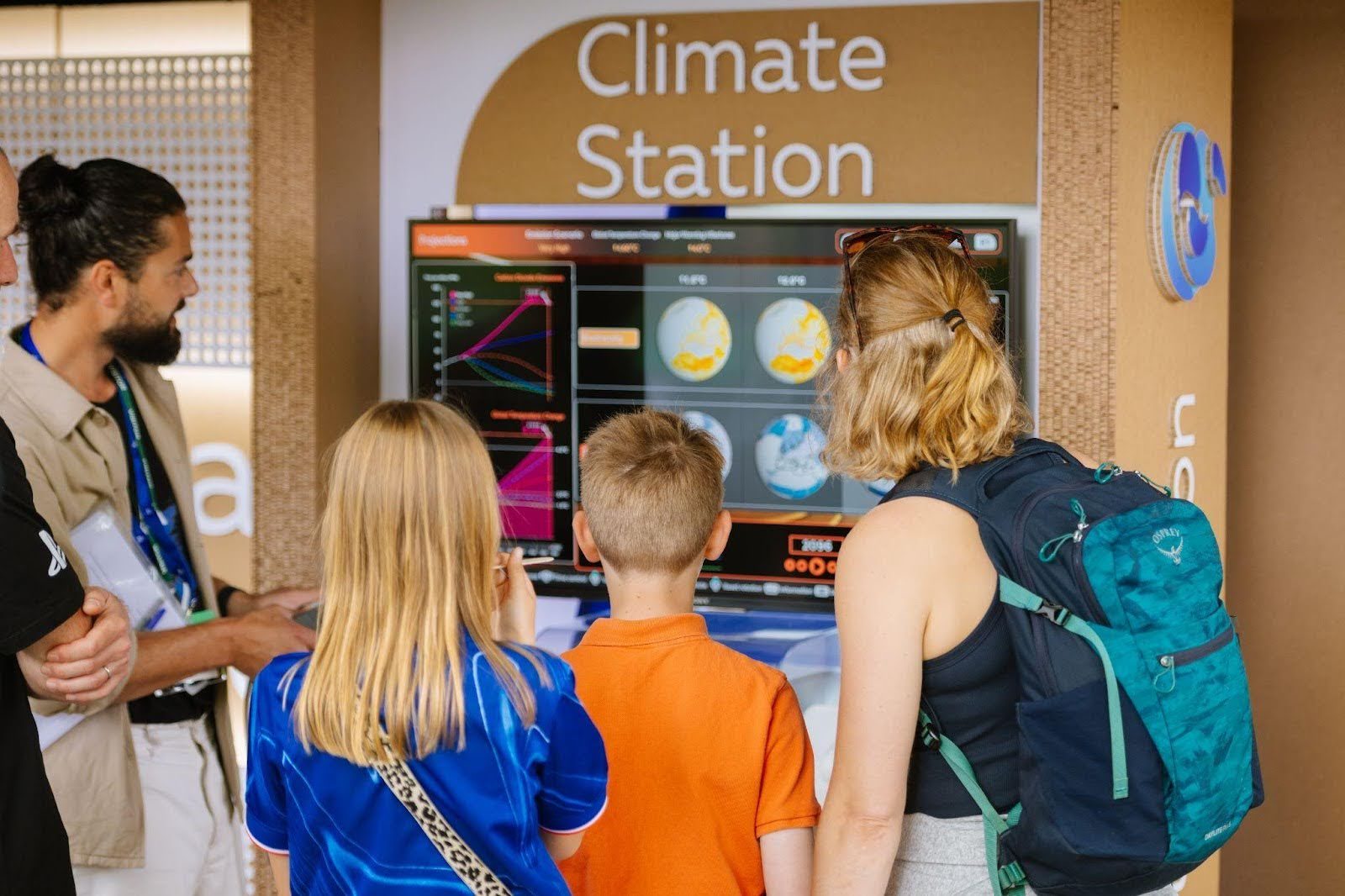 A group of children and adults gathered around a display of the Climate Station app. The screen is displaying charts and images of a globe.