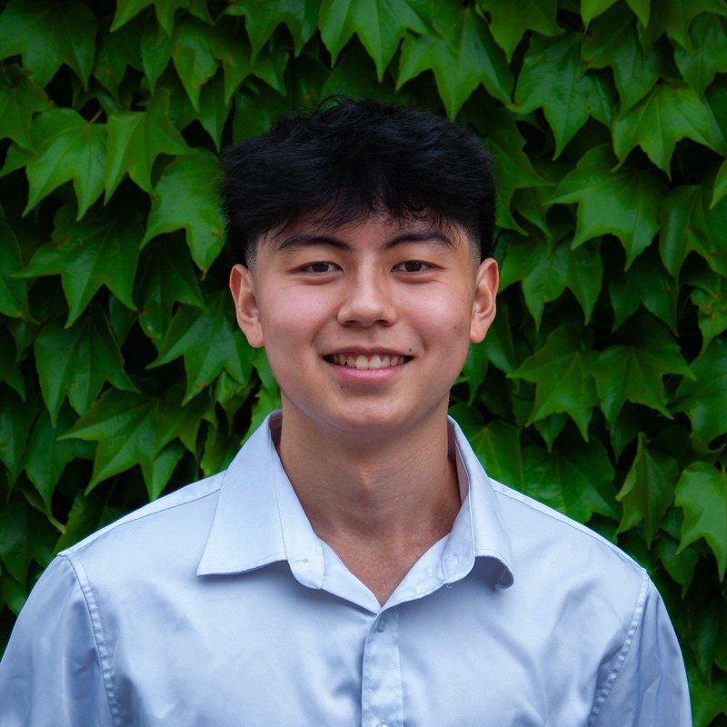 A headshot of a man with short black hair and a white shirt on a backdrop of green leaves